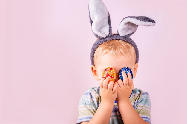 Menino bonito com orelhas de coelho na cabeça, fechando os olhos com ovos coloridos no fundo rosa.