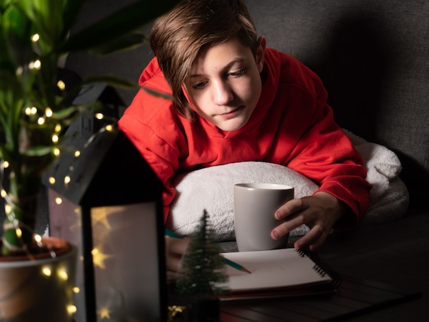 Menino bonito com uma xícara deitada no sofá e desenhando uma imagem Noite, luzes e decorações de Natal