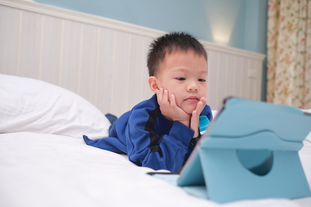 Menino bonito criança asiática deitada de bruços durante o jogo, assistindo desenhos animados, usando o computador tablet pc, crianças viciadas em gadgets, Aprendendo Tablet para crianças, conceito de brinquedo educacional de criança