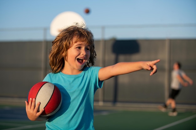 Menino bonito criança joga basquete, apontando para cima mostrando o gesto. Crianças ativas desfrutando de jogos ao ar livre com bola de basquete.