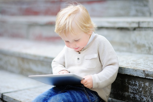 Menino bonito da criança loira brincando com um tablet digital ao ar livre