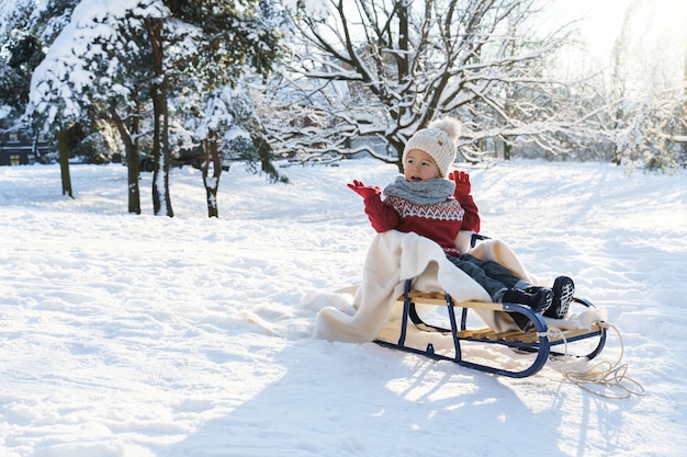 Menino bonito da criança sentado no trenó em um parque da cidade de neve durante o dia ensolarado de inverno