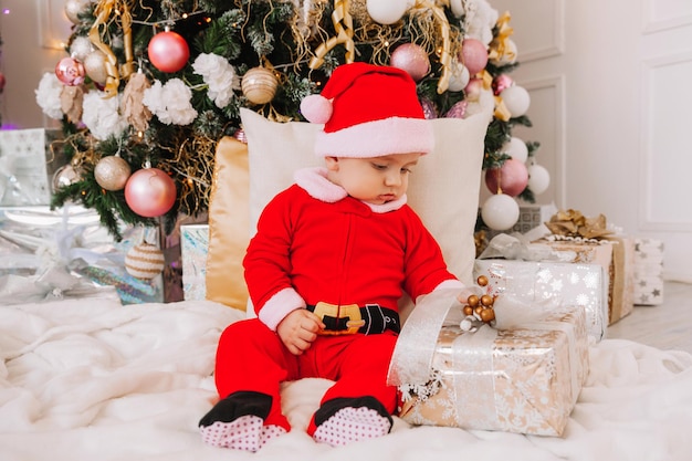 Menino bonito de 6 meses de idade, vestido de Papai Noel, senta-se perto da árvore de Natal. O bebê está sentado no chão com presentes perto da árvore de Natal. Natal de 2022