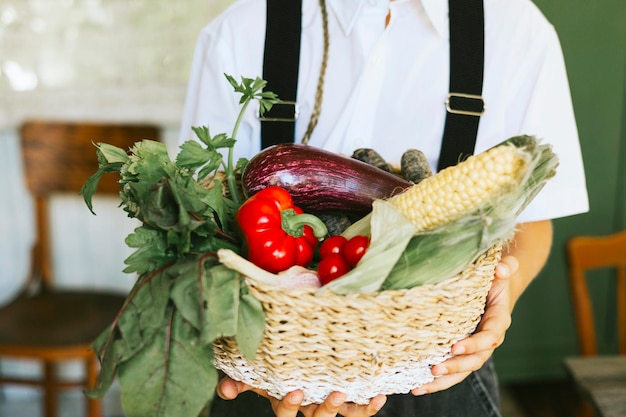 Menino bonito do fazendeiro em roupas casuais fica na varanda da casa da aldeia do país com cesta de legumes comida ecológica da colheita de outono do jardim