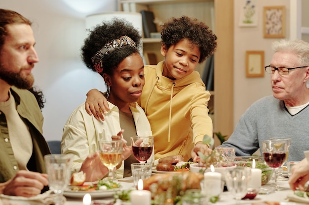 Menino bonito e afetuoso levando comida da mesa festiva e abraçando sua mãe