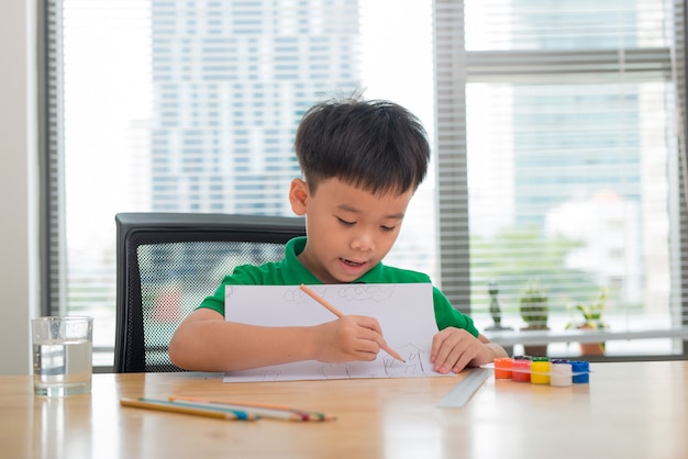 Menino bonito e confuso e sorridente fazendo lição de casa, colorir, escrever e pintar. Crianças pintam. Crianças desenham. Pré-escolar com livros na biblioteca. Lápis coloridos e papel sobre uma mesa. Menino criativo.