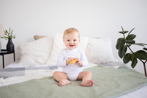 Menino bonito e saudável em um quarto espaçoso e brilhante está sentado em uma cama na roupa de cama branca, a criança está sorrindo têxteis para o quarto e o berçário