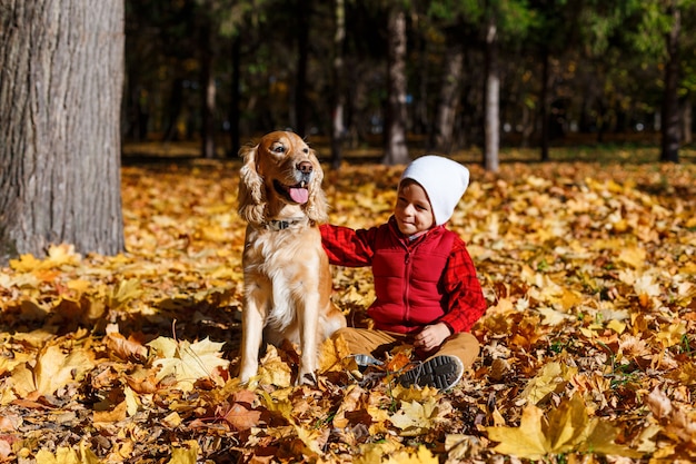 Menino bonito, feliz, branco de camisa vermelha, sorrindo e brincando com o cachorro entre as folhas amarelas. Criança se divertindo no parque outono. Conceito de amizade entre crianças e animais de estimação, família feliz