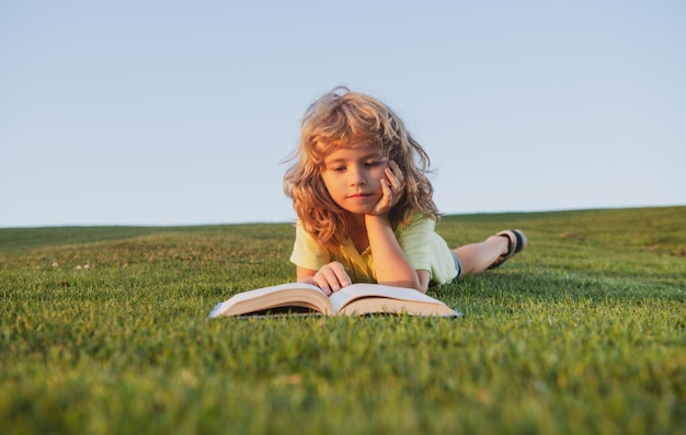 Menino bonito lendo livro ao ar livre no campo de grama verde criança inteligente lendo livro deitado na grama su