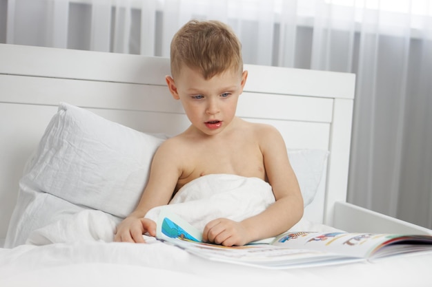 Menino bonito lendo um livro em uma cama branca