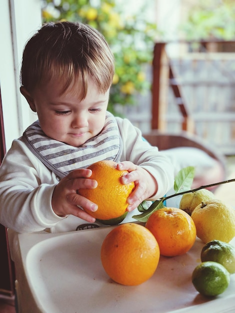 Foto menino bonito segurando frutas de laranja enquanto está sentado em casa