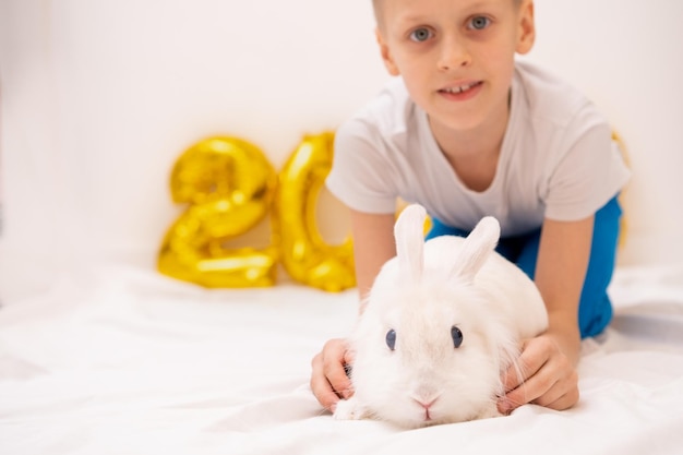 Menino bonito sorri com coelho branco closeup em um fundo branco com balões de folha de ouro Coelhinho da Páscoa o ano do coelho Ano Novo 2023 Natal Amor pelos animais