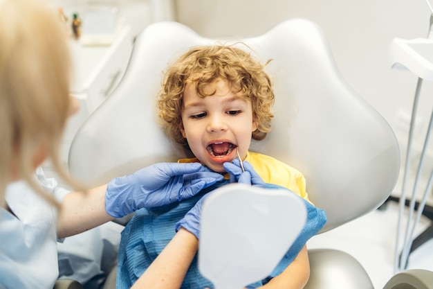 Menino bonito visitando o dentista, tendo seus dentes verificados por dentista no consultório odontológico.