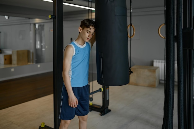 Menino cansado de pé perto do saco de boxe descansando depois de um duro treino