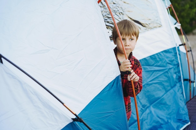 Menino caucasiano bonitinho olhando para fora da tenda turística Conceito de acampamento familiar
