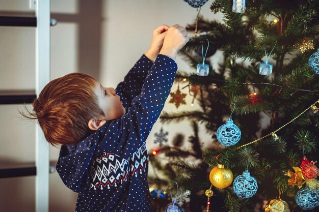 Menino caucasiano bonito decorando a árvore de Natal com decorações cintilantes