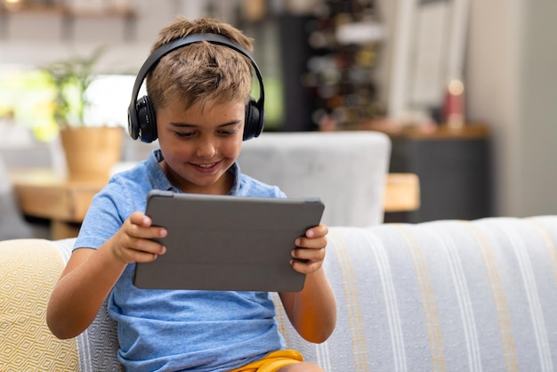 Menino caucasiano bonito usando fones de ouvido e assistindo vídeo em um tablet digital enquanto está sentado no sofá