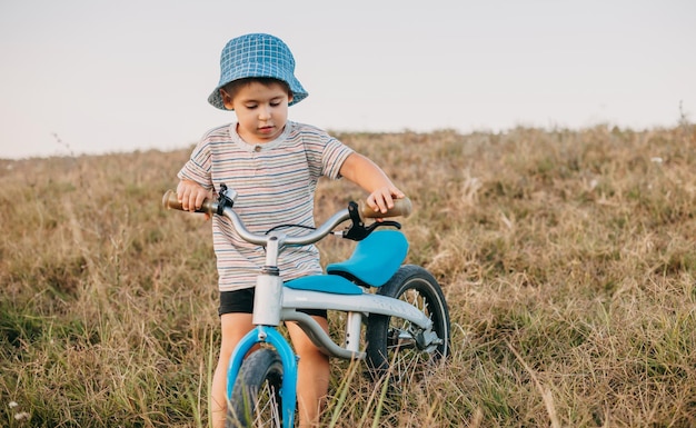 Menino caucasiano com chapéu azul segurando a bicicleta no campo, recreação de verão