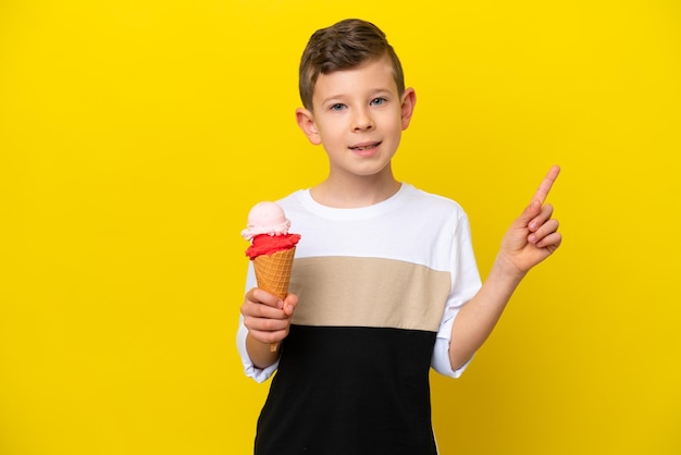 Menino caucasiano com um sorvete de corneta isolado em fundo amarelo mostrando e levantando um dedo em sinal do melhor