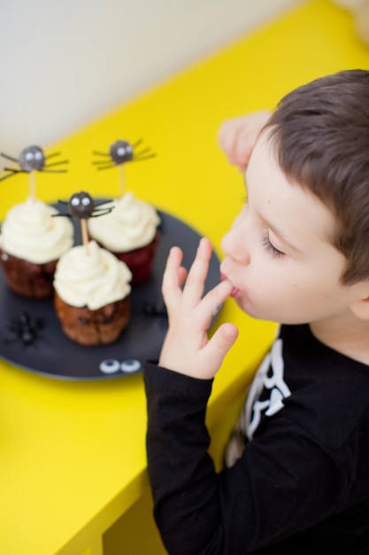 Menino caucasiano criança come muffins ou cupcakes de Halloween com creme branco e decorações de aranhas Comida saborosa e assustadora na festa de Halloween