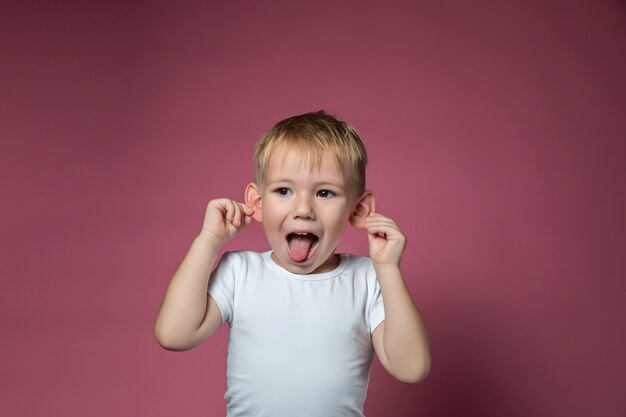 Menino caucasiano de 3 anos fazendo caretas, mostrando a língua, olhando para a câmera no fundo rosa