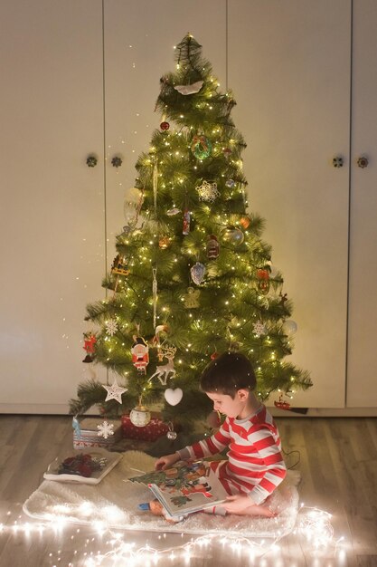 Menino caucasiano de pijama listrado com livro sentado perto da árvore de natal interior acolhedor de natal com decorações e luzes