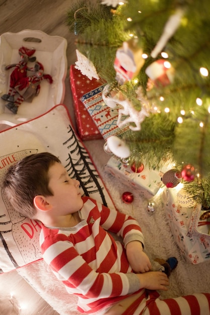 Menino caucasiano de pijama listrado dormindo sob a árvore de Natal Interior acolhedor de Natal com decorações e luzes