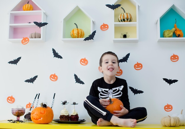 Menino caucasiano infantil em traje de esqueleto segura abóbora curvada e senta-se na mesa com comida e decorações de Halloween