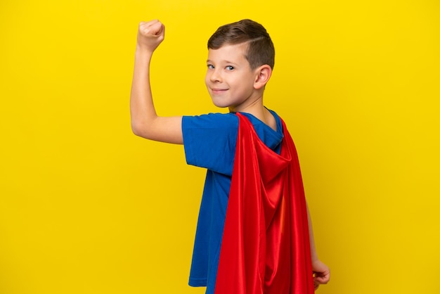 Menino caucasiano isolado em fundo amarelo em traje de super-herói e fazendo um gesto forte
