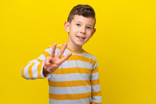 Menino caucasiano isolado em fundo amarelo feliz e contando três com os dedos