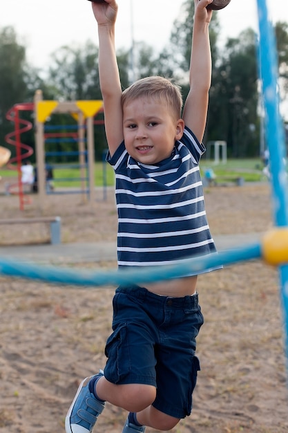 Menino caucasiano pendurado na barra de macacos pela mão para fazer exercício no playground ao ar livre.