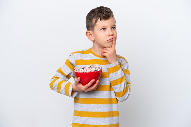 Menino caucasiano segurando uma tigela de cereal isolada no fundo branco com dúvidas ao olhar para cima