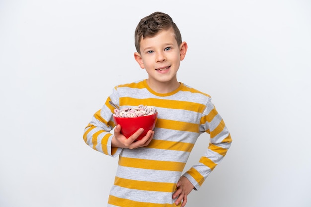 Menino caucasiano segurando uma tigela de cereal isolada no fundo branco posando com os braços no quadril e sorrindo