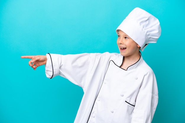 Menino chef isolado em fundo azul, apontando o dedo para o lado e apresentando um produto