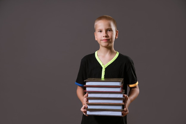 Menino colegial com camiseta preta segurando uma pilha de livros em uma parede cinza