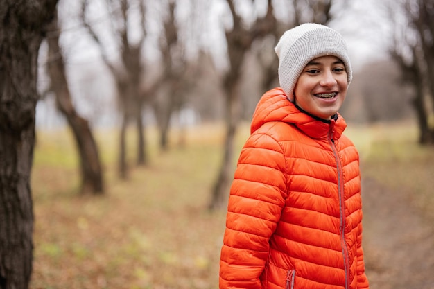 Menino com aparelho usa jaqueta laranja e chapéu branco na floresta de outono