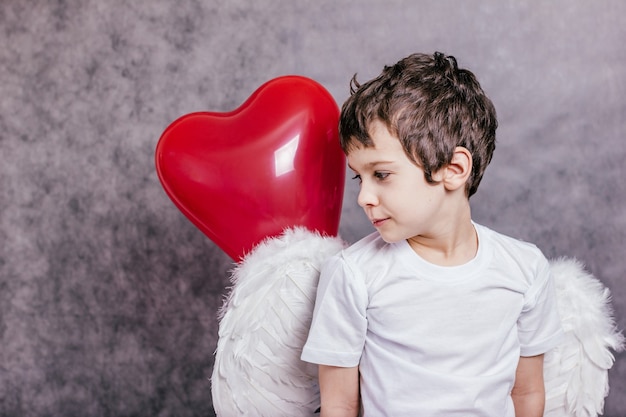 Foto menino com asas de anjo escondido atrás de uma bola vermelha em forma de coração