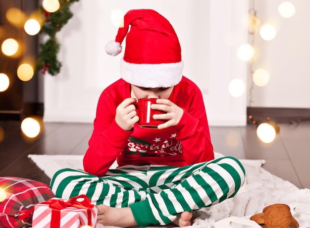 Menino com boné de Papai Noel vermelho engraçado e pijama de Natal sentado em uma manta e bebendo chocolate