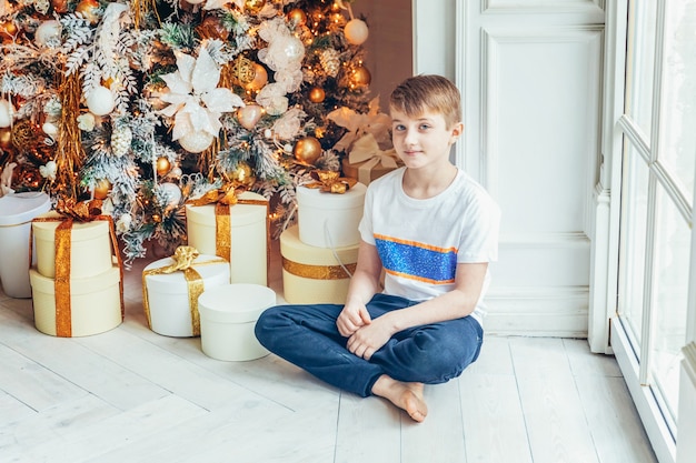 Menino com caixa de presente perto de árvore de Natal na véspera de Natal em casa. Criança em uma sala iluminada com decoração de inverno. Família feliz em casa. Tempo de dezembro de ano novo de Natal para o conceito de celebração.