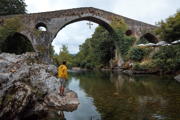 menino com capa de chuva amarela próximo ao rio Sella olhando para a bela ponte romana de Cangas de Onis, nas Astúrias