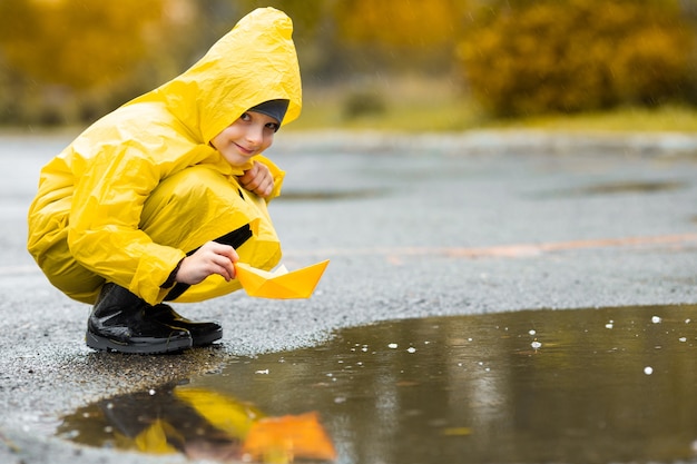 Menino com capa impermeável amarela e botas de borracha pretas, brincando com o brinquedo do barco feito à mão de papel em uma poça ao ar livre na chuva no outono.