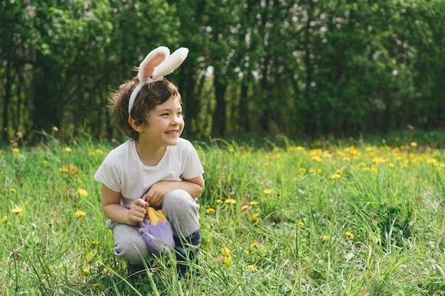 Menino com cesto de ovos e orelhas de coelho em caça de ovos de Páscoa no jardim ensolarado da primavera