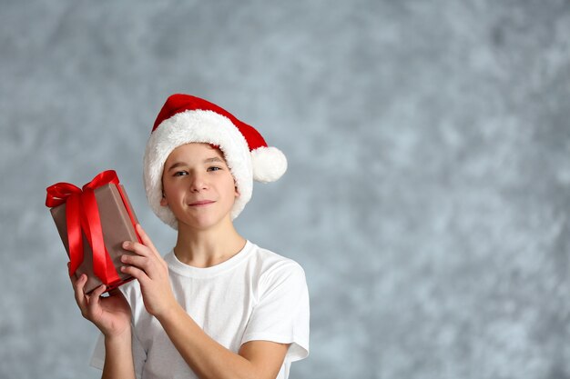 Menino com chapéu de Papai Noel com caixa de presente na parede cinza