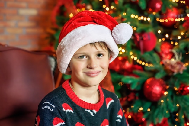 Menino com chapéu de Papai Noel em casa perto da árvore de Natal. Criança feliz esperando o natal