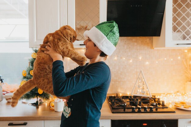 Menino com chapéu de Papai Noel segura um gato ruivo na cozinha. Natal. férias de inverno. Belo retrato de um menino com um gato ruivo nos braços.