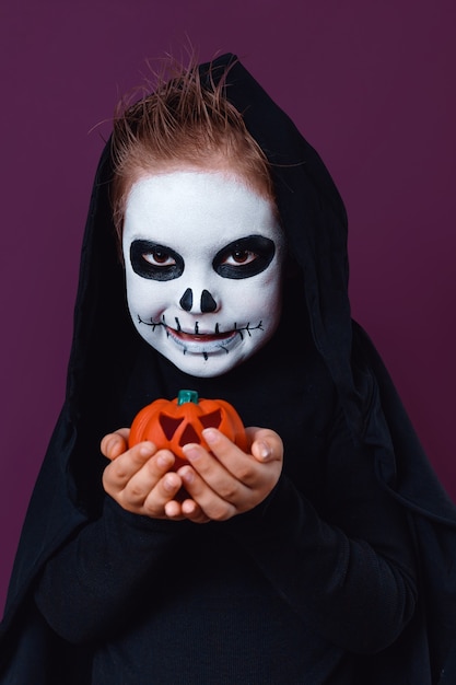 Menino com fantasia de halloween detém abóbora Jack-o-lantern em fundo roxo. Maquiagem de Halloween.