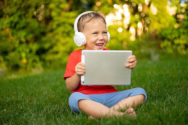 Menino com fones de ouvido sem fio, segurando um computador tablet ao ar livre em um parque de verão, sorrindo