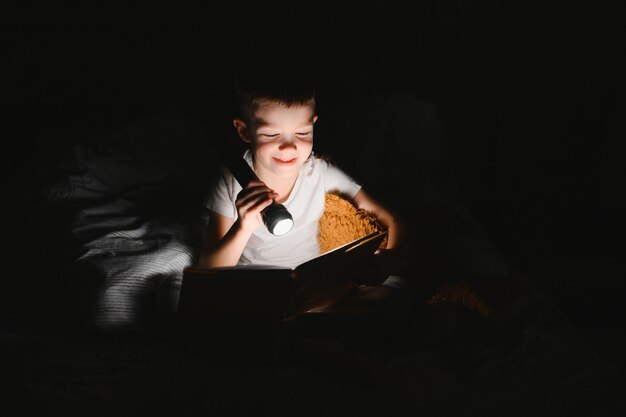 Menino com lanterna lendo livro debaixo do cobertor em casa