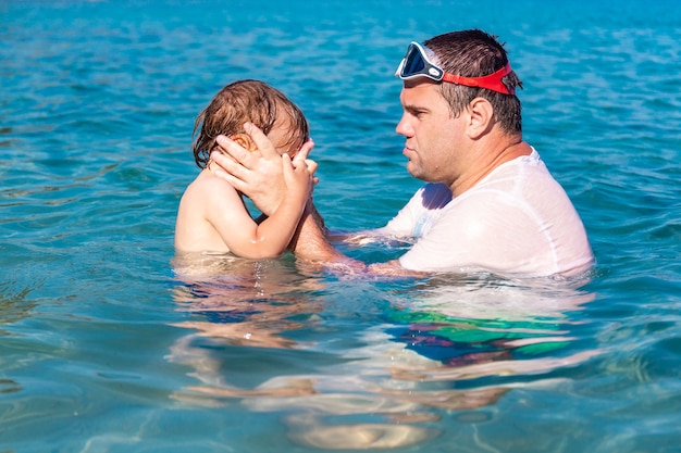 Menino com medo de olhos ardentes de água no mar. Pai e filho nadando e brincando juntos na água do mar nas férias de verão