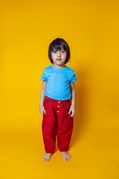 Menino com roupas coloridas, calça vermelha e camiseta azul, parado em uma parede amarela